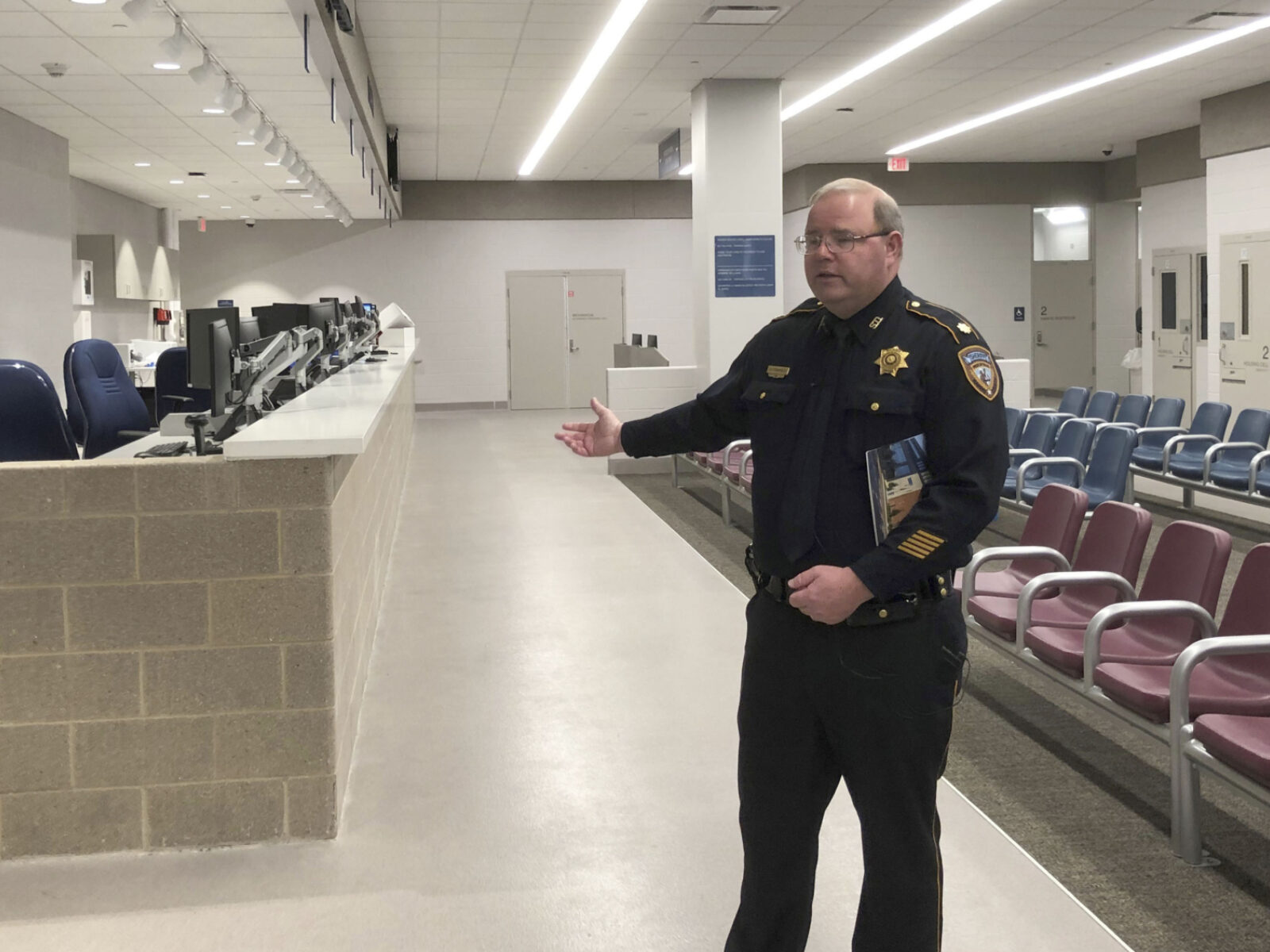 A sheriff's office major stands in a brightly lit intake center with waiting chairs and a cinderblock counter lined with computers.