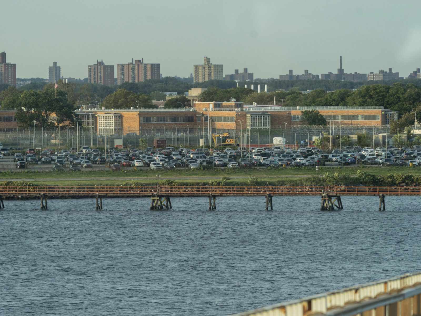 A view of Rikers Island jail complex from afar shows body of water in front, then fencing and buiildings.