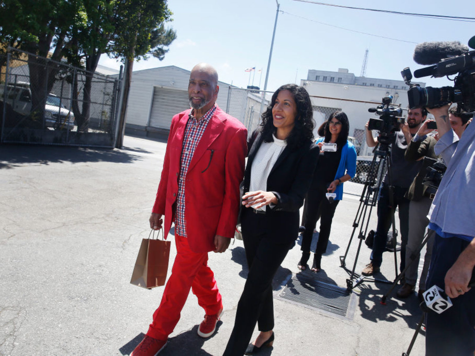 Kenneth Humphrey in red suit and carrying briefcase, and Anita Nabha walk outside with media cameras following them.