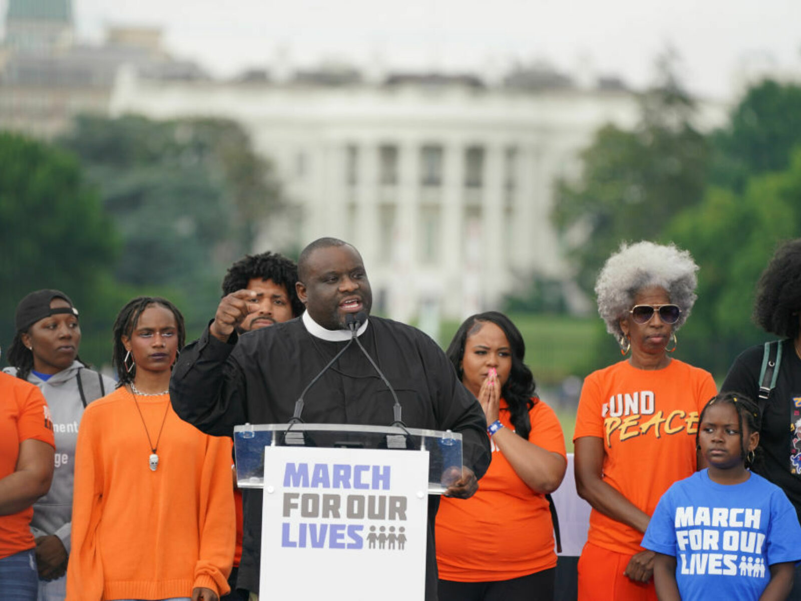 Pastor Mike McBride stabds at a podium speaking passionately and flanked by people young and old on both sides as the White House is seen in the background.