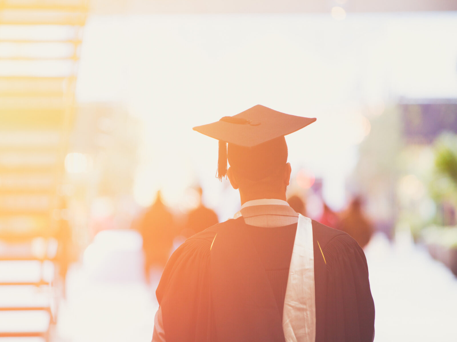 Graduate in cap and gown walks away in street in sun dappled scene.