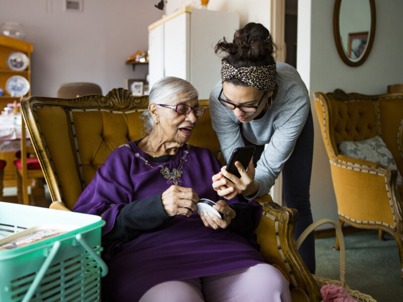 An elderly woman sits in a chair at home looking at her phone while a young woman leans over to help her.