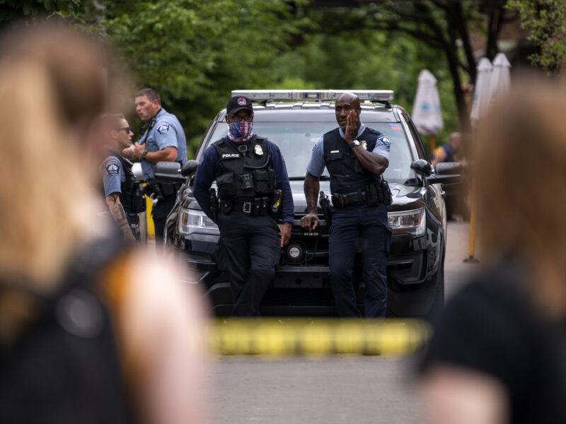 Officers stand in front of patrol vehicle while onlookers stand nearby.