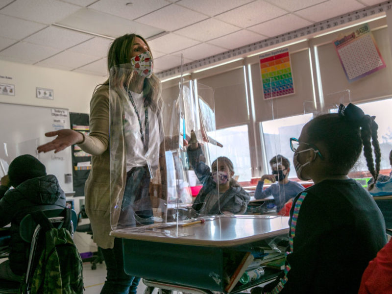 Teacher wearing mask stands at front of classroom with students sitting at desks.