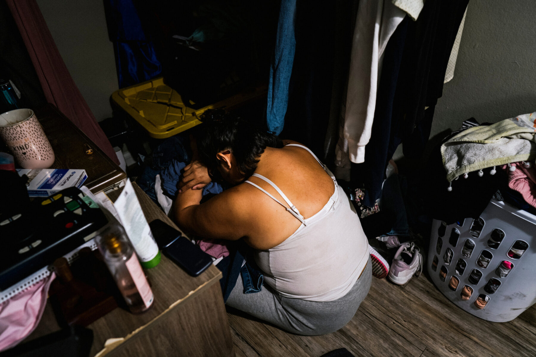 Melissa kneels on the floor resting her head on a pile of clothes in a closet. Clothes hang on one side of her body. On the other side, personal items like perfumes, a candle, and a reader are seen on a dresser.